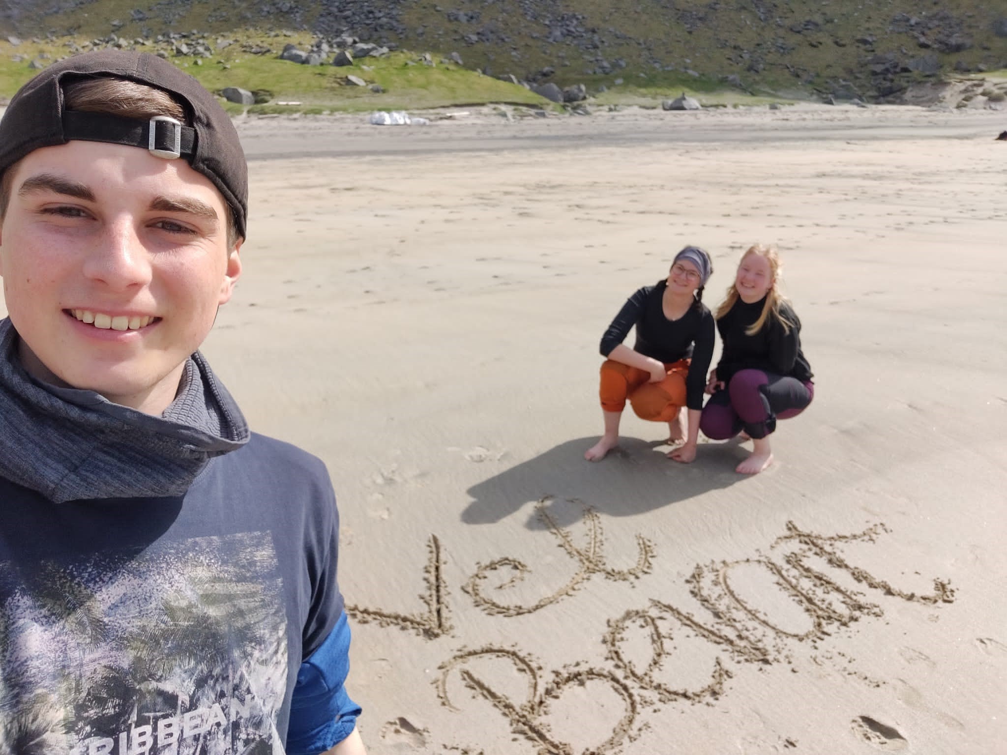 Tre personer på ei strand. Det er skrevet tekst i sanda. Foto 